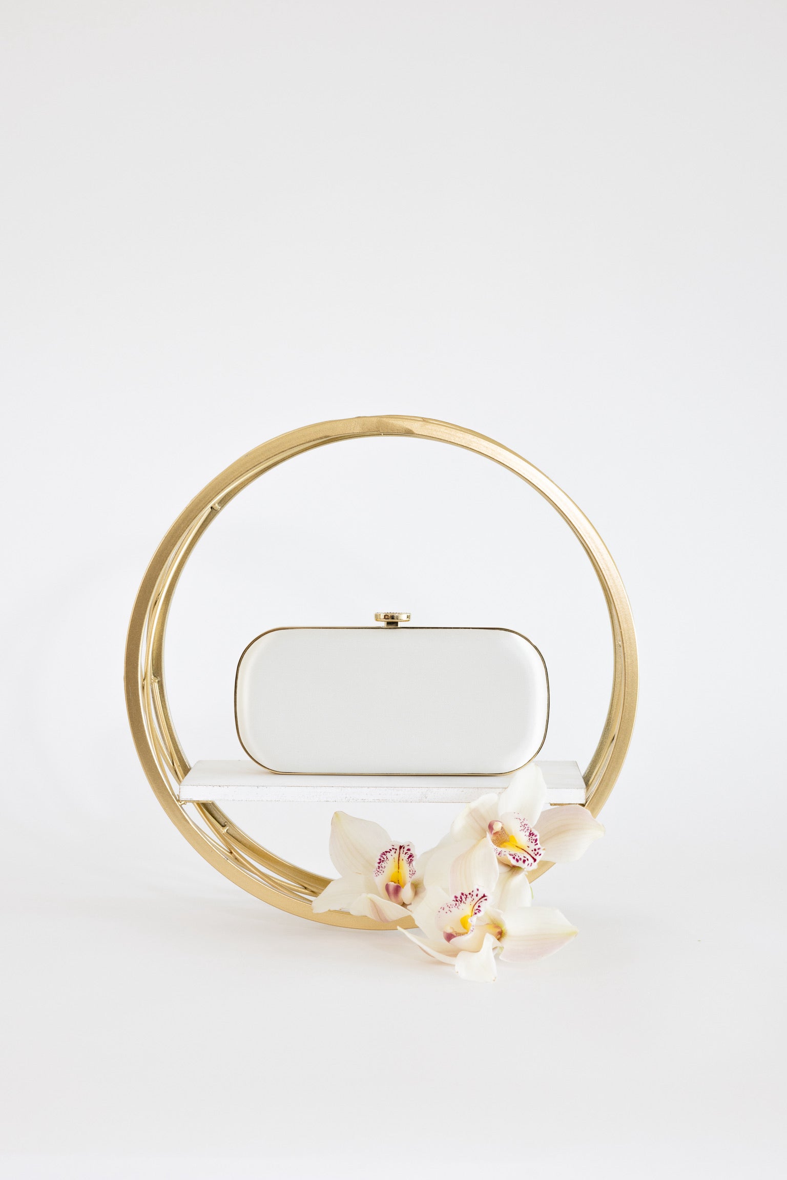 A white clutch purse displayed within a golden circular frame, accented by white orchids to the front, against a plain white background.