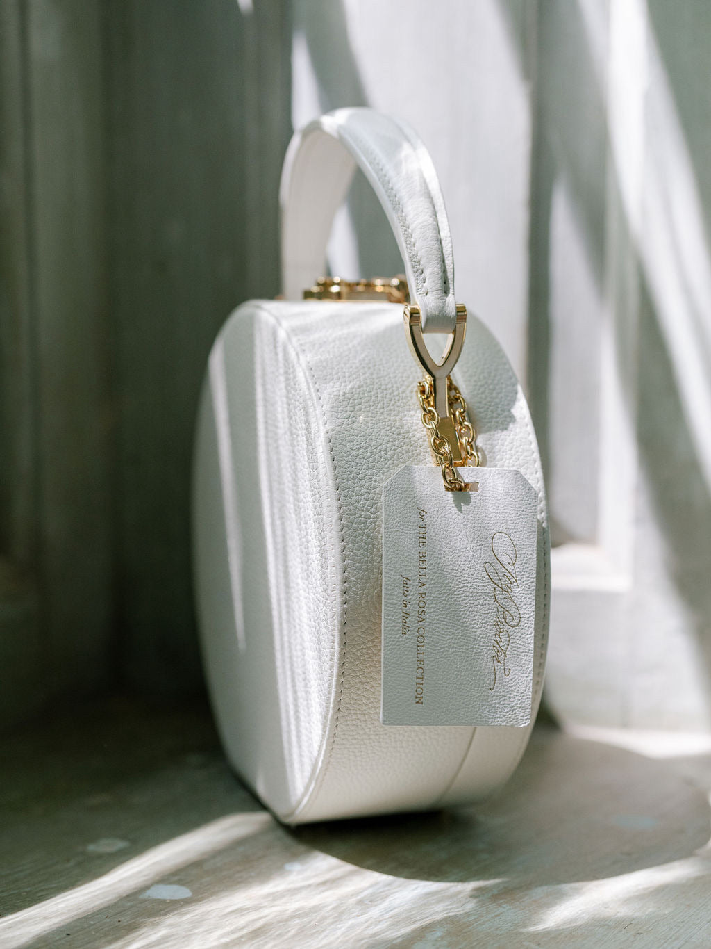 An elegant white designer handbag with a gold chain detail, placed against a light-dappled backdrop.