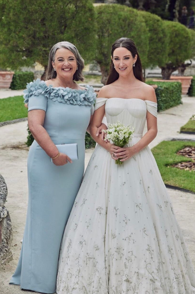 Two women dressed in formal attire, one in a blue dress with ruffle details and the other in an off-the-shoulder bridal gown with floral embellishments, posing smilingly in a garden setting.