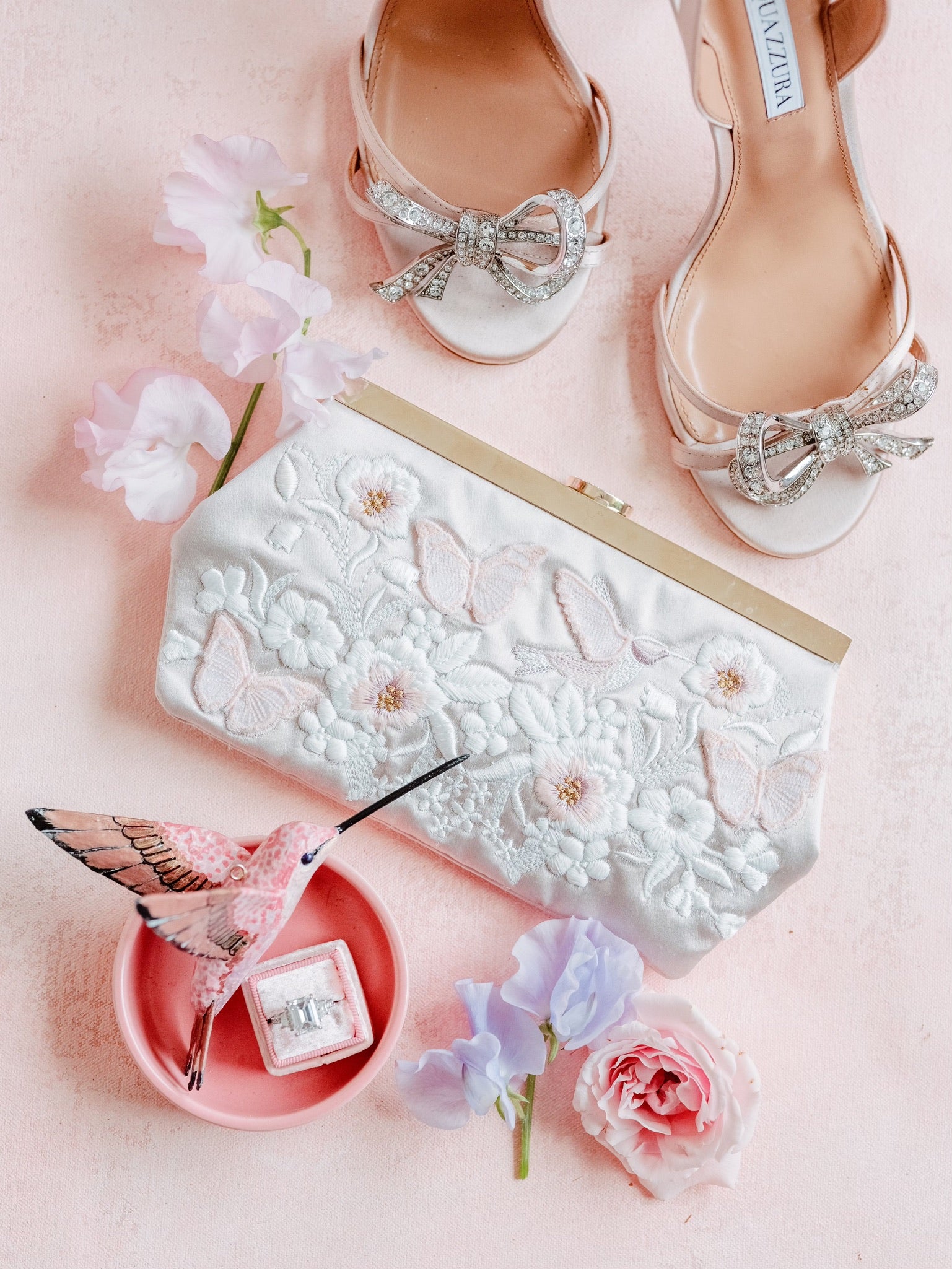 An elegant bridal arrangement featuring shimmering shoes, a floral clutch, and fresh flowers on a pink background.