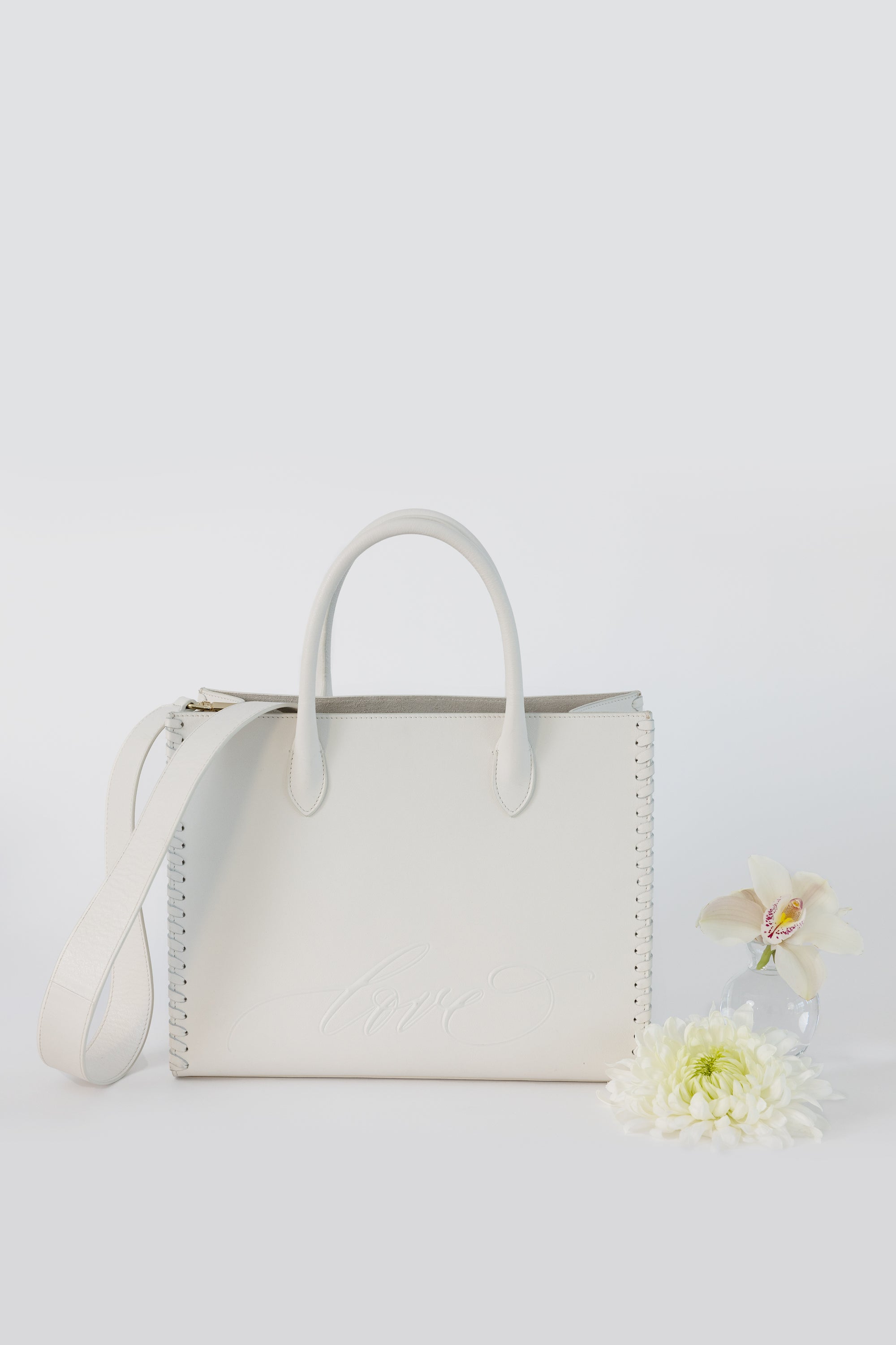 A LOVE Tote - White from The Bella Rosa Collection, featuring a shoulder strap and elegantly embossed with the word "love," is placed next to a white chrysanthemum and a light-colored orchid in a glass vase. This handbag is perfect for carrying wedding day essentials.