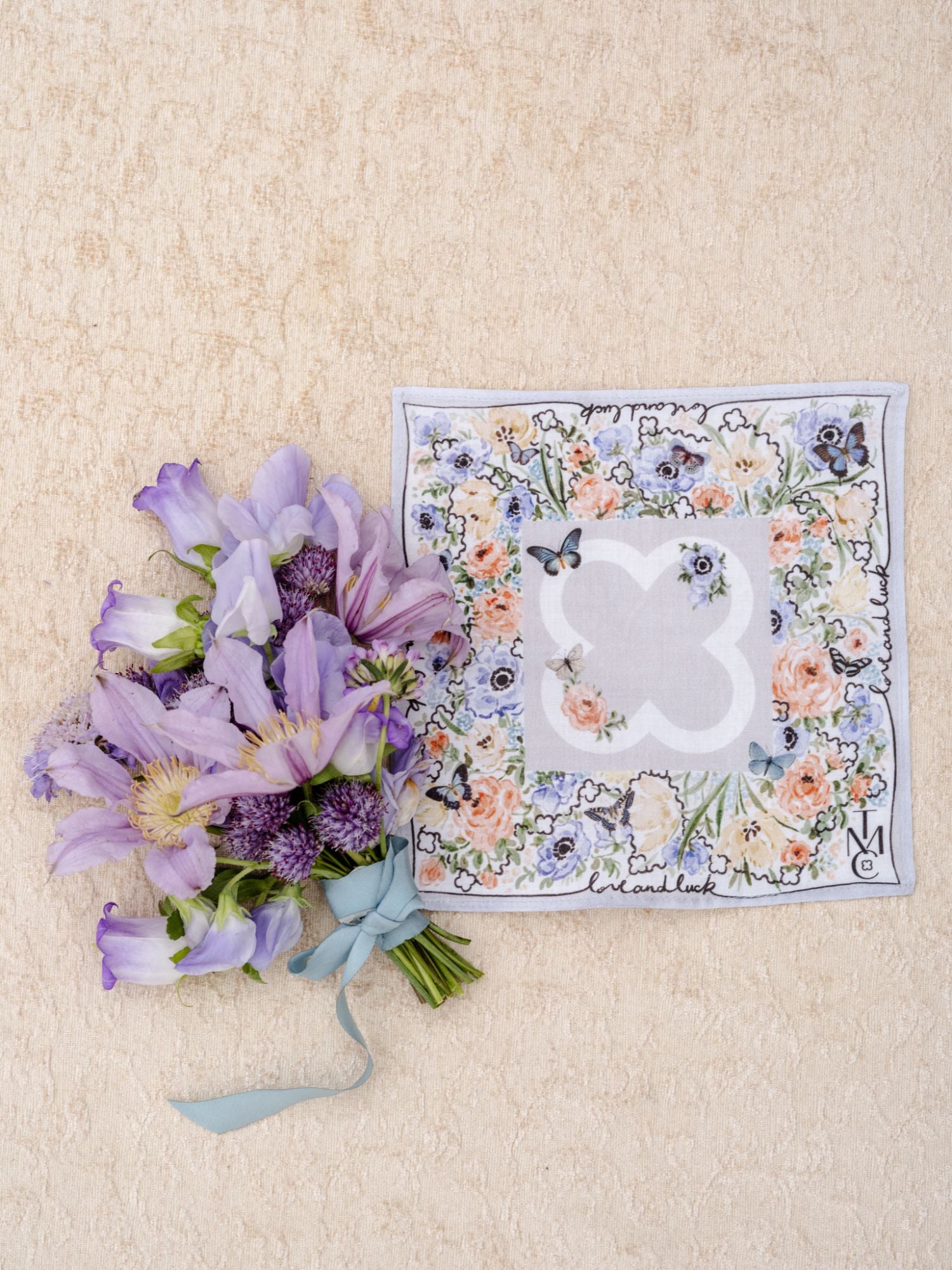 A small bouquet of purple flowers with a blue ribbon sits next to The Bella Rosa Collection Wedding Floral Handkerchief, displayed on a beige textured surface.