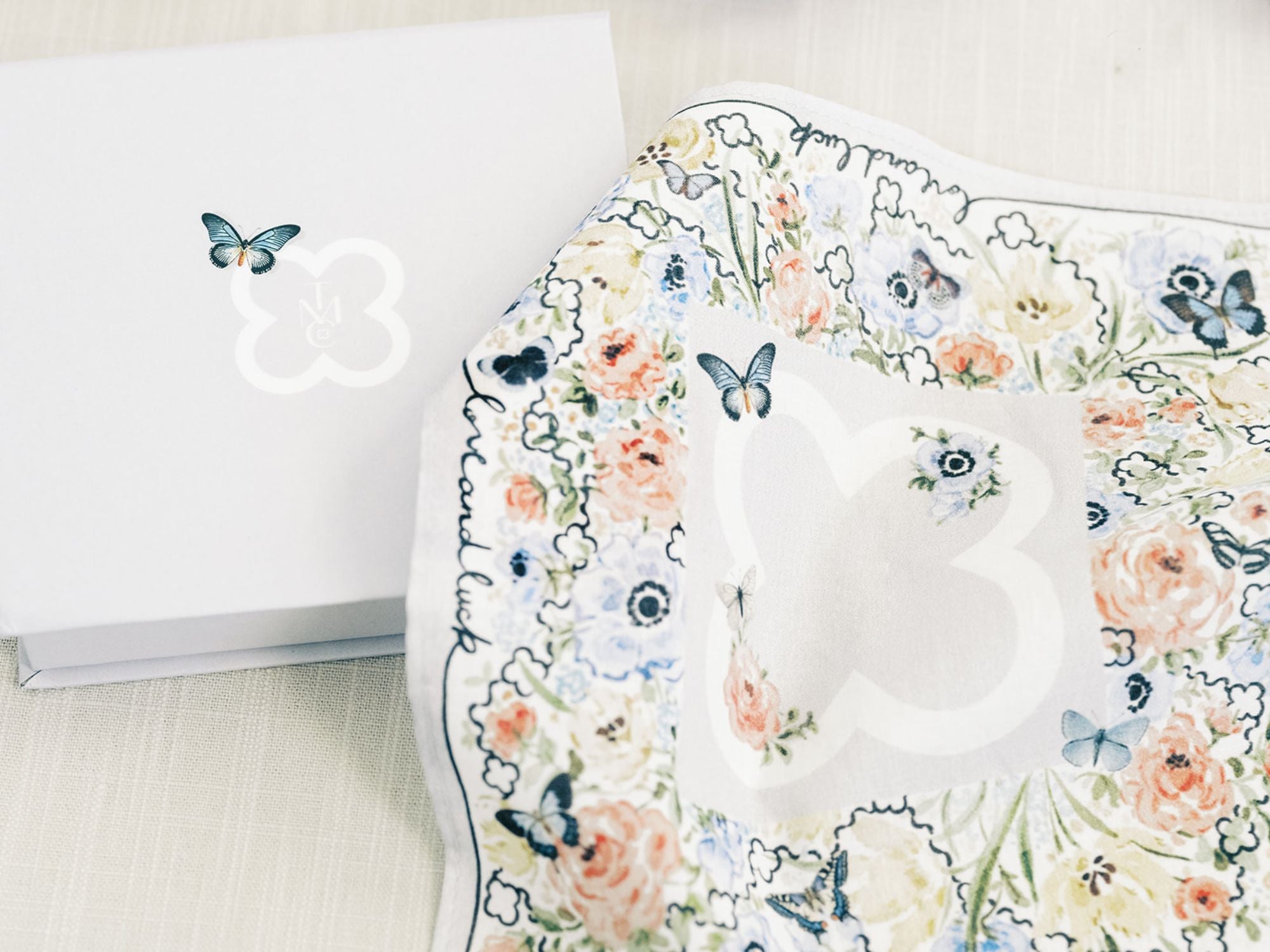 The Bella Rosa Collection’s Wedding Floral Handkerchief—an Italian cotton piece with butterfly motifs—comes in a white box with butterfly logo, set on light fabric, making it an elegant luxury bridal gift.