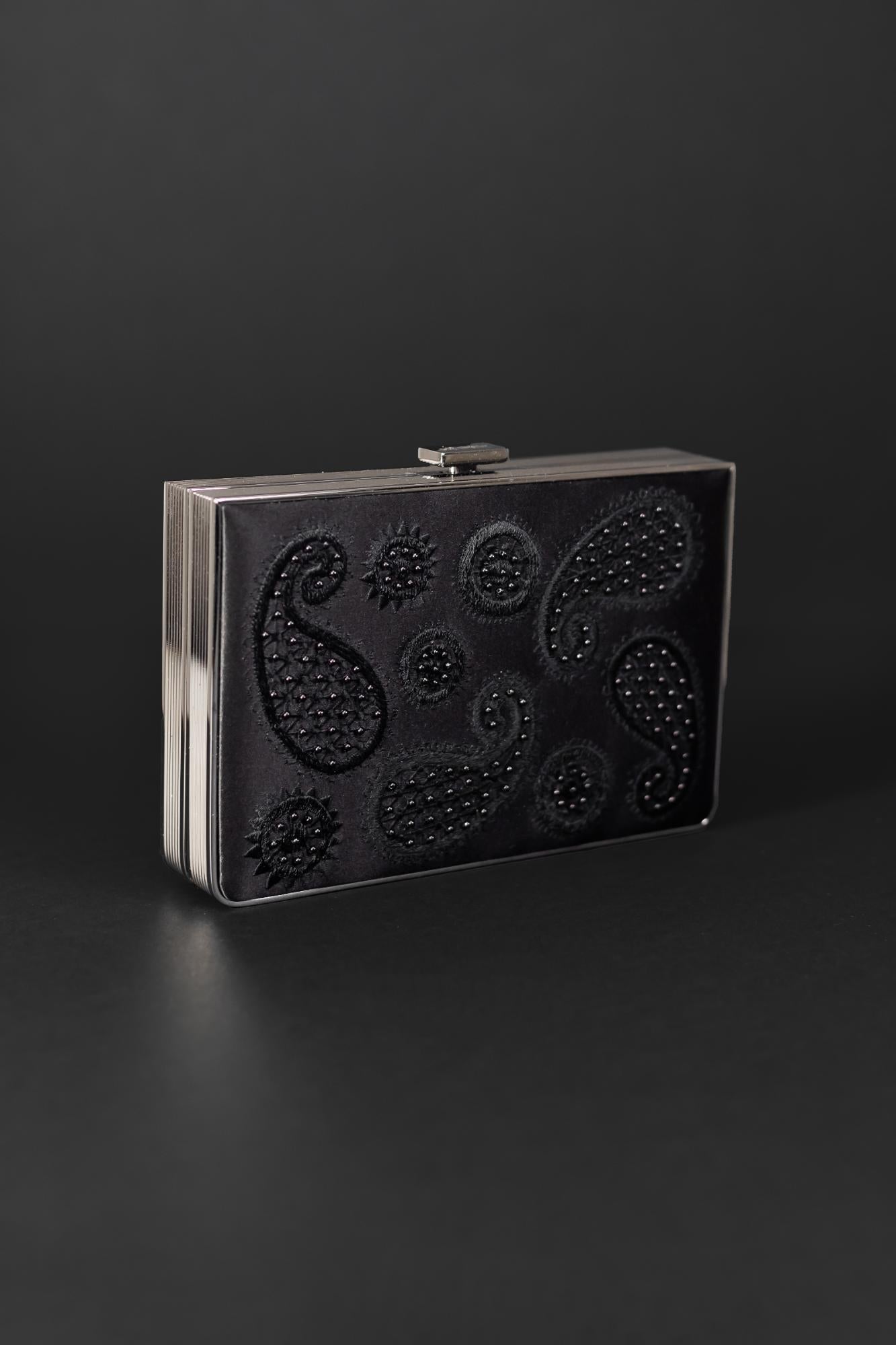 The Venezia Clutch x MICAELA from The Bella Rosa Collection is a sleek, rectangular accessory in black satin, featuring 3D embroidery of embossed paisley and floral patterns against a dark background.