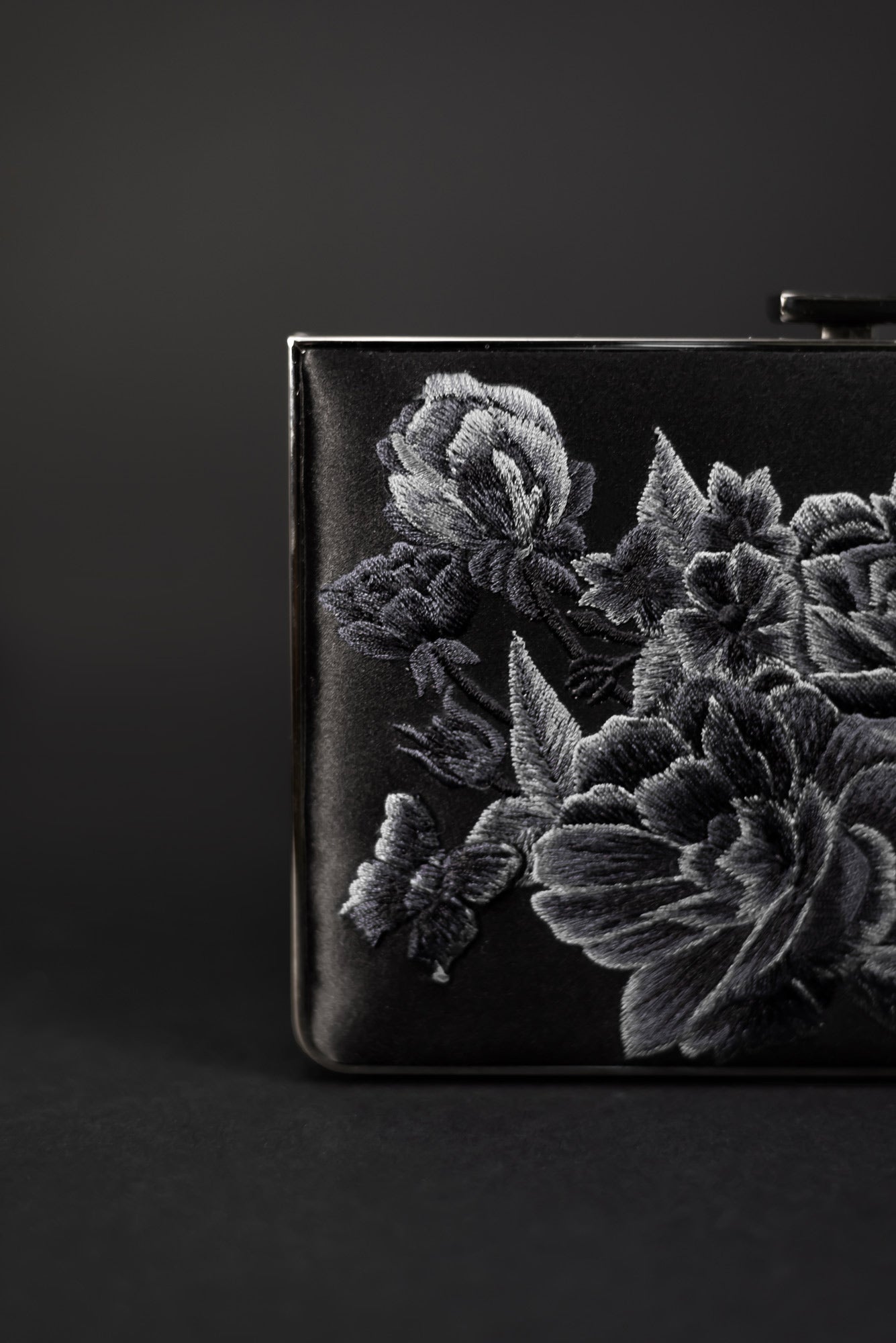 A Venezia Clutch x MICAELA from The Bella Rosa Collection, showcasing exquisite gray floral embroidery on black satin, is photographed against a dark background, highlighting its elegance. This exclusive piece is part of our sample sale.
