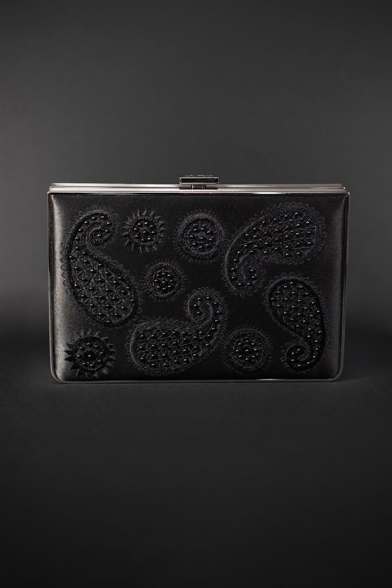 Description: The Bella Rosa Collection's Venezia Clutch x MICAELA Black Satin Paisley Embroidery - Sample Sale features a sleek black, rectangular design adorned with intricate paisley and floral patterns embossed on its surface, enhanced by exquisite 3D embroidery against the dark background.