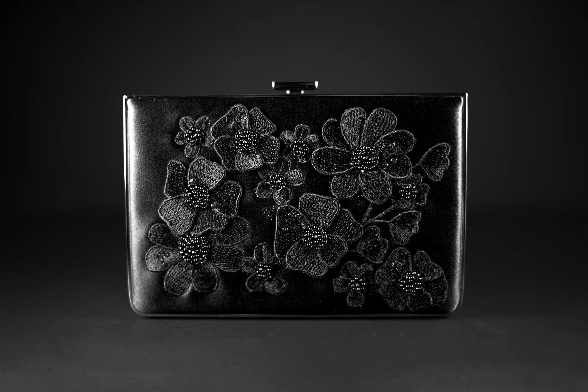 The Venezia Clutch x MICAELA Black Satin Floral Embroidery from The Bella Rosa Collection, a sample sale item, features intricate 3D floral embroidery, making it a stunning accessory against the black background.