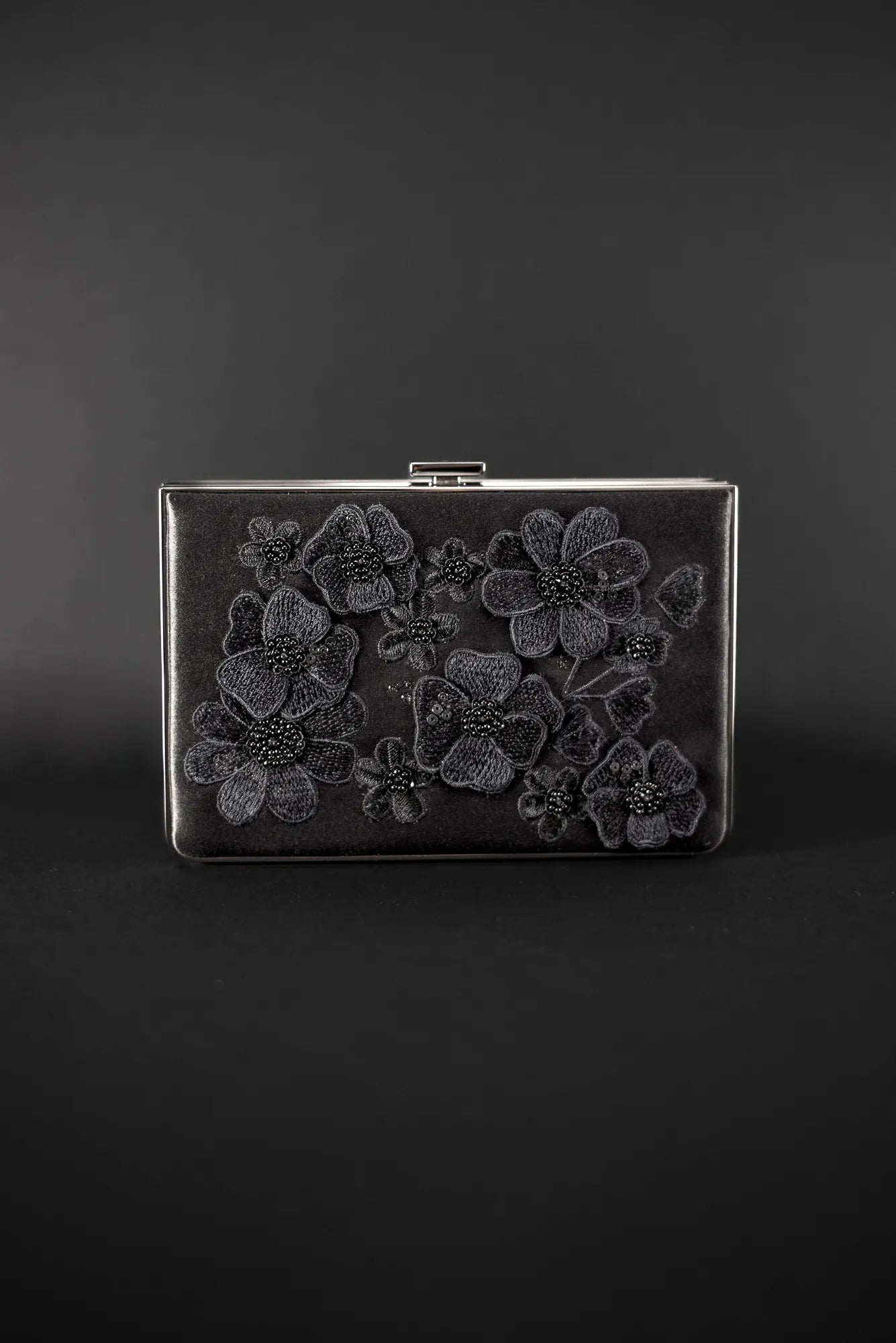 The Venezia Clutch x MICAELA Black Satin Floral Embroidery from The Bella Rosa Collection, a rectangular black purse adorned with 3D floral embroidery, is showcased against a dark background. Sample Sale item.