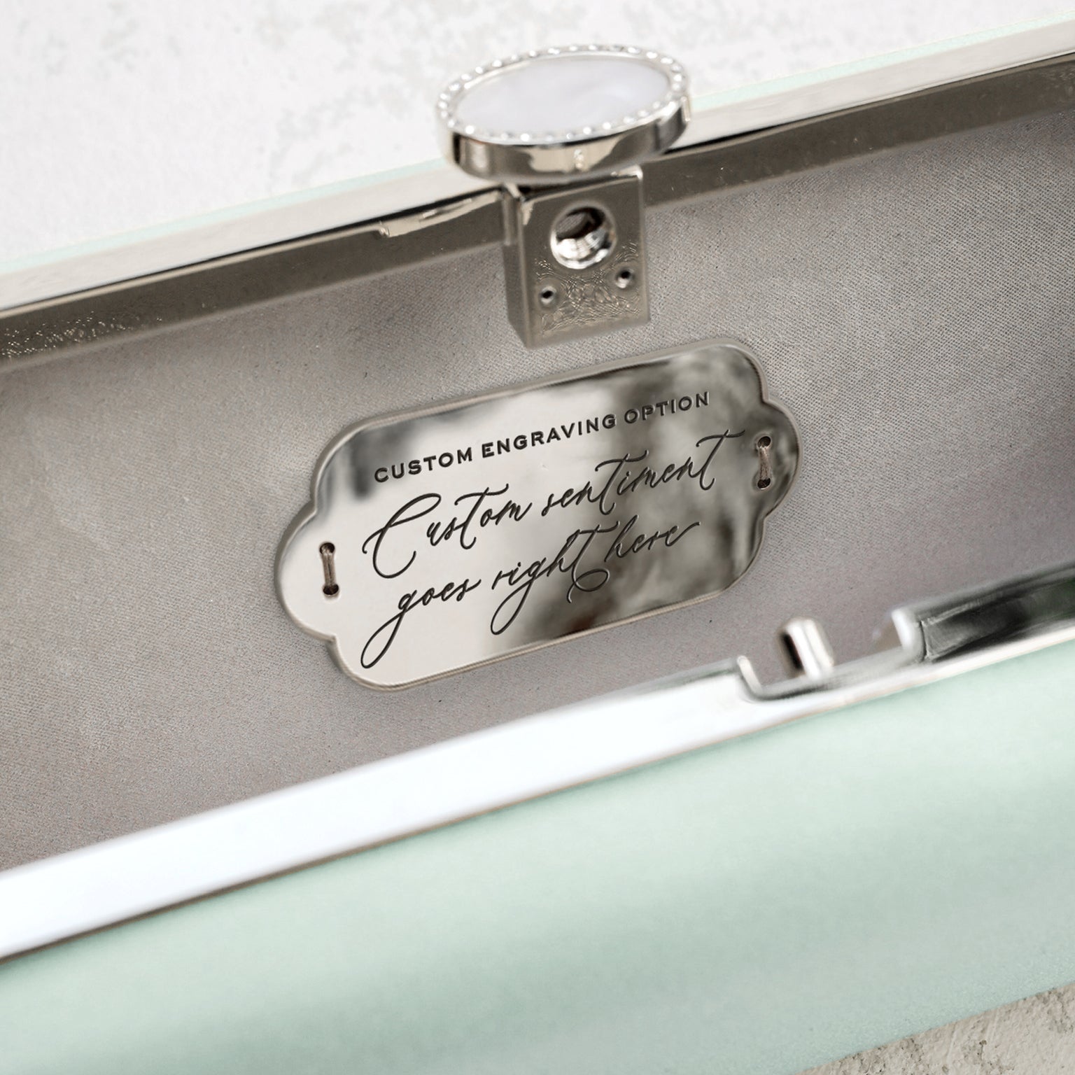 An open jewelry box with a customizable engraving plate on the inside of the lid, suggesting a personalized message can be added for The Bella Rosa Collection&#39;s Bella Clutch Sage Green Satin Petite.