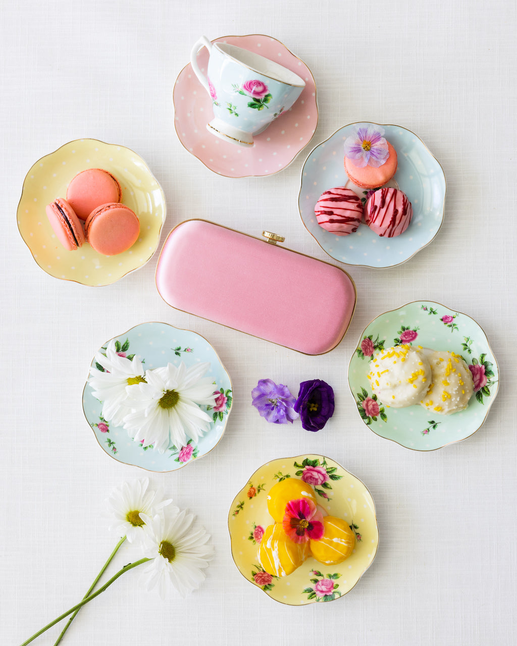 An array of pastel-colored plates with assorted desserts, a Bella Clutch Pink Petite from The Bella Rosa Collection, and fresh flowers on a light background.