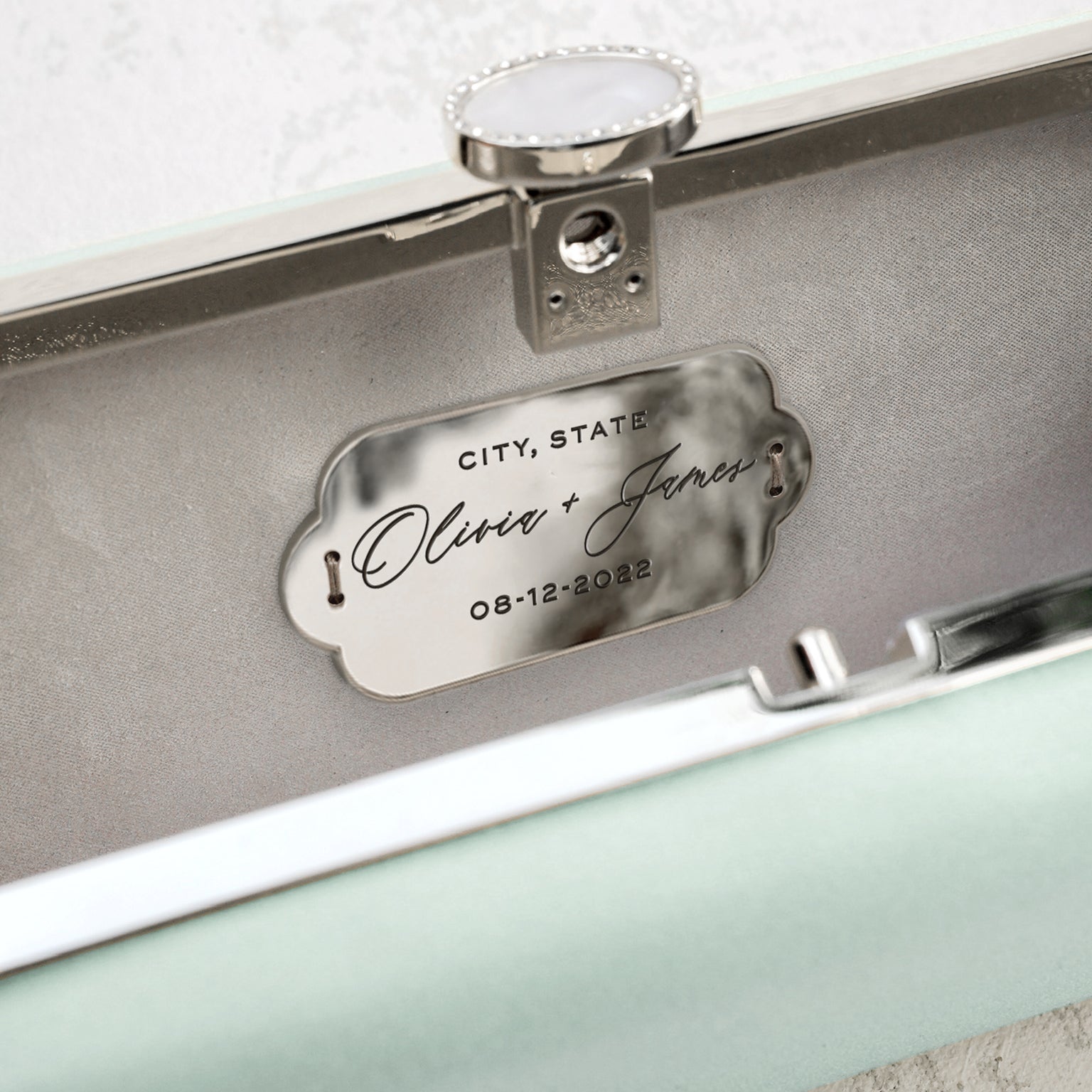 Custom-engraved plaque with the names &#39;Olivia &amp; James&#39; and the date &#39;08-12-2022&#39; inside a Sage Green Bella Clutch Satin Petitie from The Bella Rosa Collection.