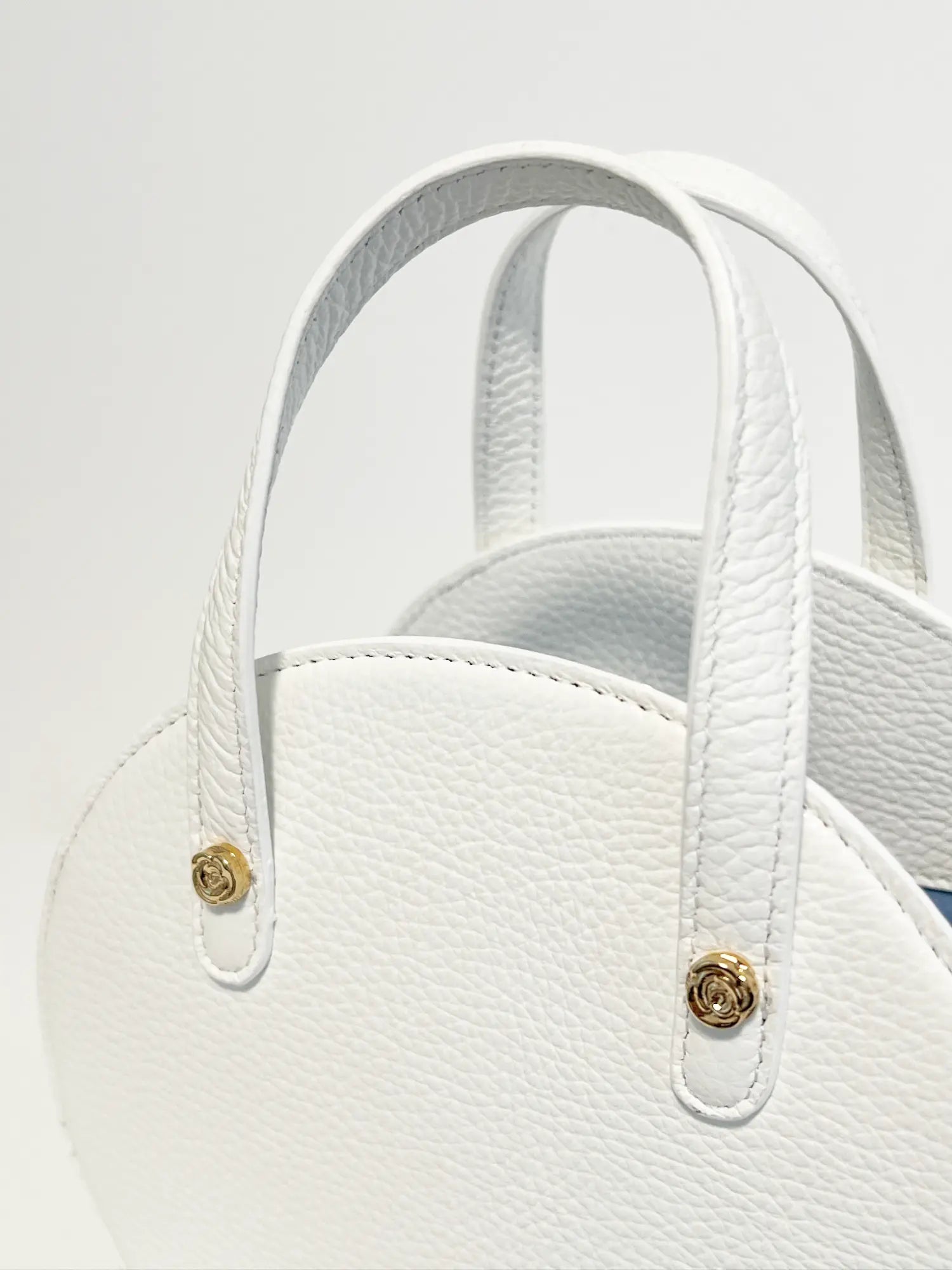 Close-up of a white textured Harper Hat Box Handbag White Leather handbag with two handles and golden metallic clasps, presented against a light gray background from The Bella Rosa Collection.