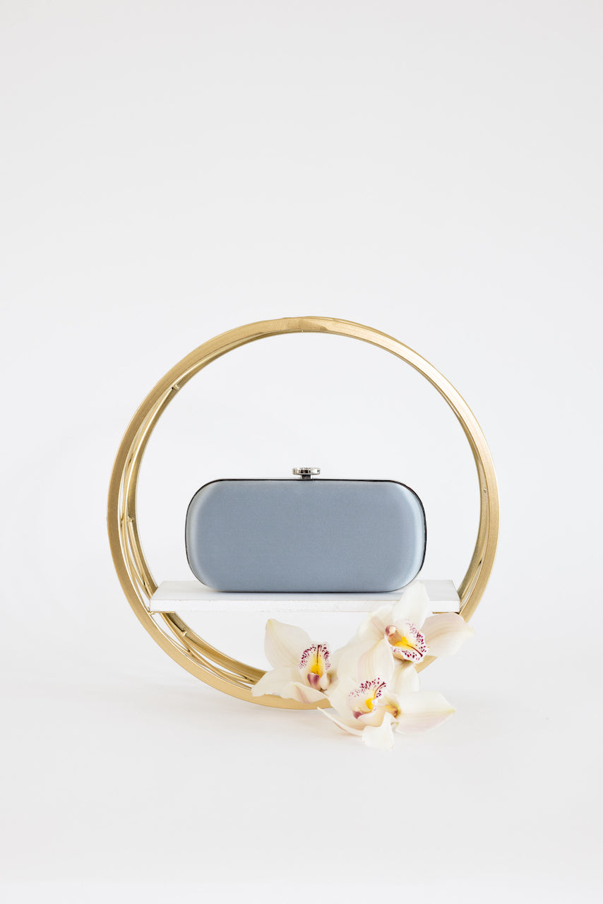 A Bella Clutch Steel Blue Satin Grande evening bag from The Bella Rosa Collection displayed with an elegant orchid flower and encircled by a gold frame on a white background.