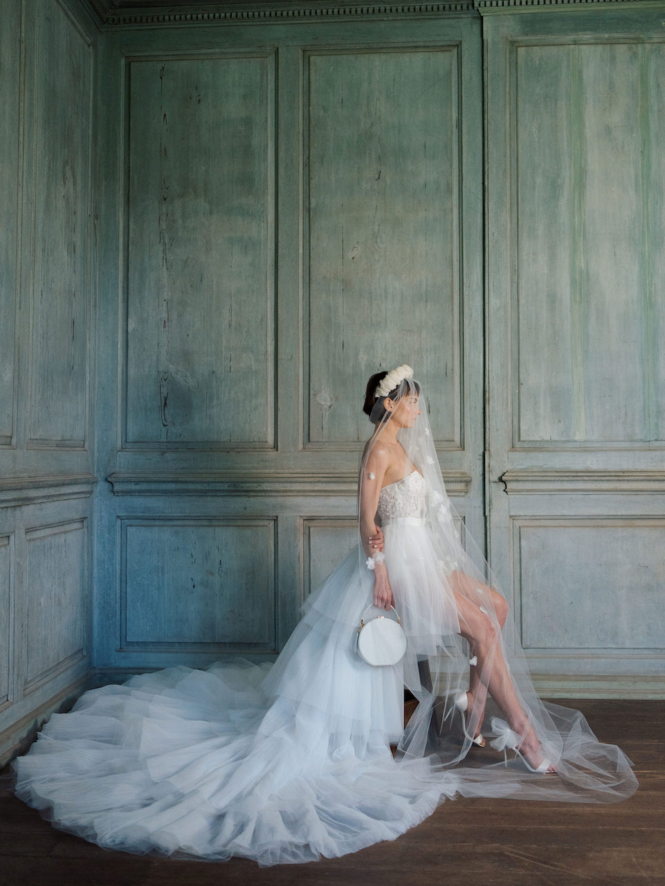 A vintage-inspired bride in a white wedding dress sitting on a wooden floor with The Bella Rosa Collection&#39;s &quot;The Original Hat Box&quot;.