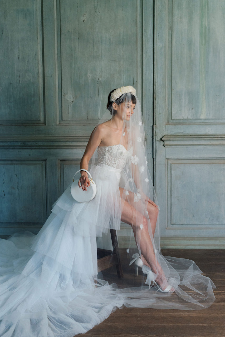 A vintage-inspired bride in a wedding dress sitting on a chair with The Bella Rosa Collection's The Original Hat Box beside her.