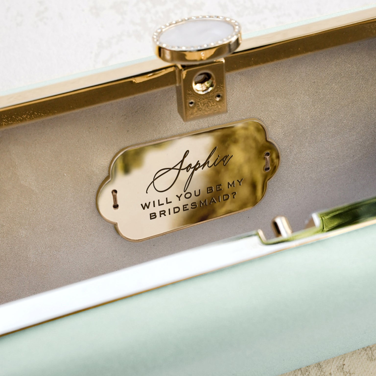 A customized Bridesmaid Proposal Box with an Engraved Bella &#39;Be My Bridesmaid&#39; Clutch Bag Gift from The Bella Rosa Collection, with a gold tag on it.