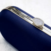 The Navy Blue Satin Bella Clutch, an elegant wedding handbag from The Bella Rosa Collection, features a blue clutch embellished with a radiant silver ring.